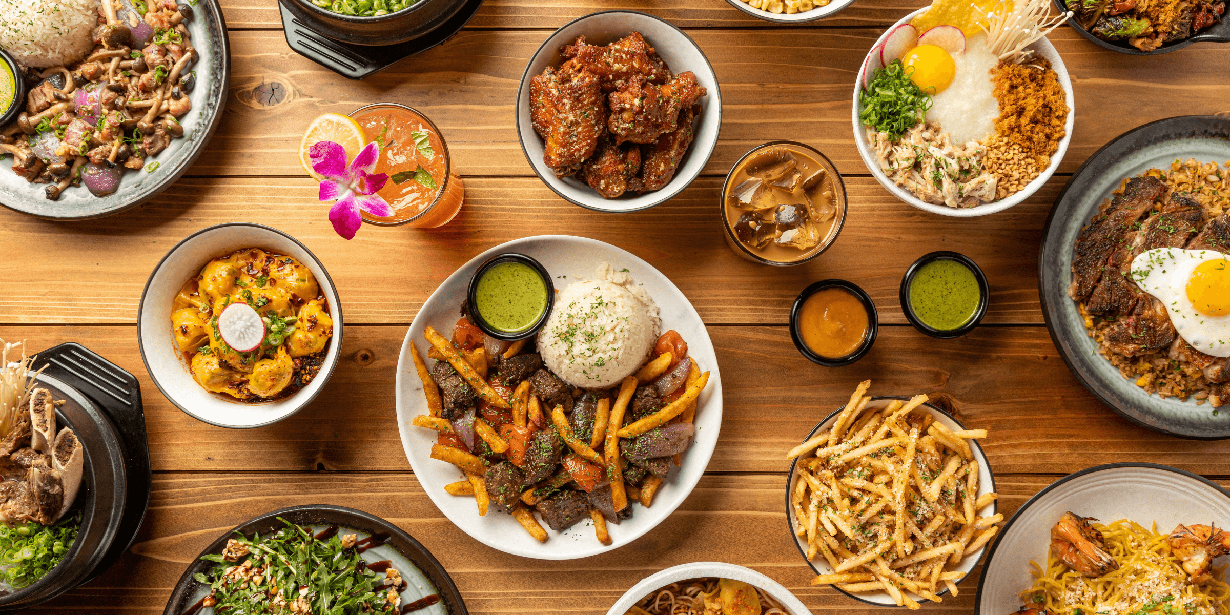 An overhead spread from VOX Kitchen featuring filet mignon lomo saltado with fries and rice, spicy wontons, parmesan garlic fries, crispy chicken wings, noodle dishes, and other signature Asian fusion plates arranged across a wooden table.