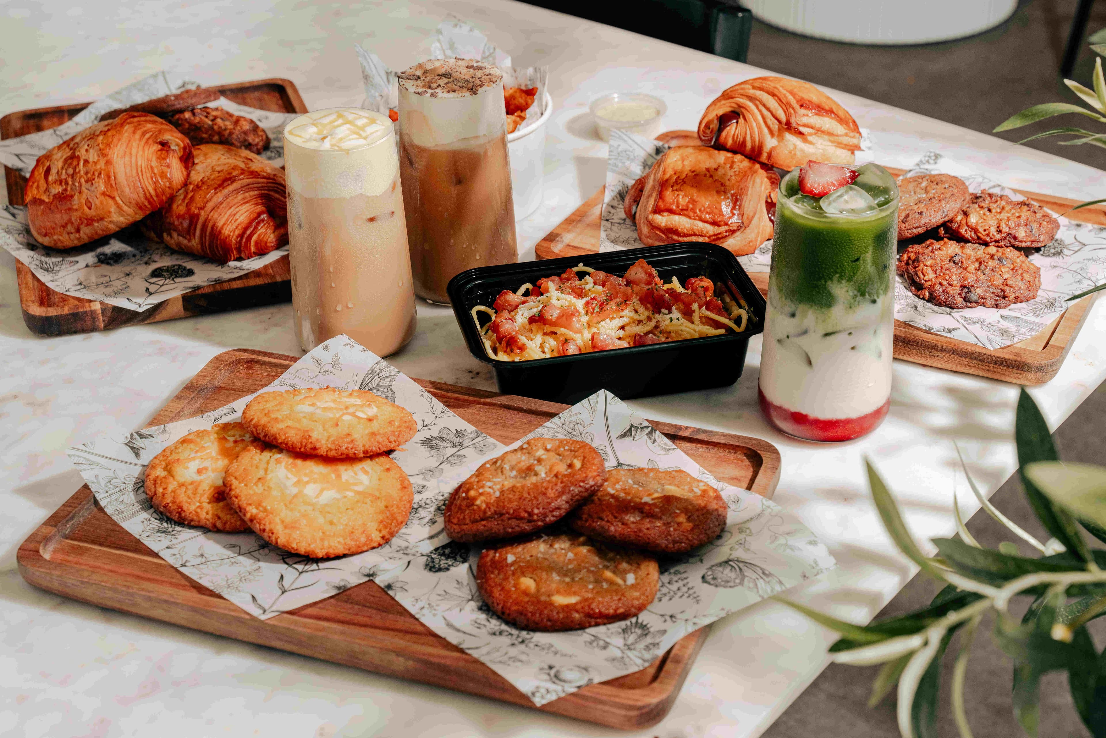 An assorted spread from Kei Coffee House featuring buttery croissants, fresh pastries, house-baked cookies, creamy pasta, and signature drinks including strawberry matcha and iced lattes arranged across a bright café table.