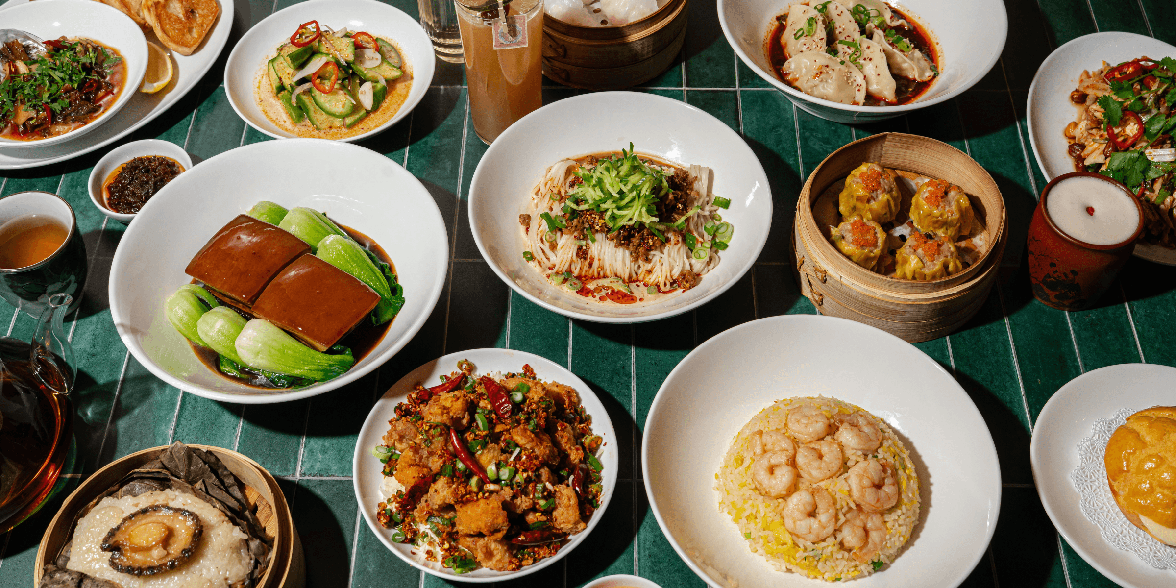 An overhead spread from QUA Contemporary Chinese Cuisine showcasing shrimp fried rice, spicy cucumber salad, shumai, chili wontons, chicken cartilage with mala BBQ spice, abalone sticky rice, braised Dong Po pork belly, and dan dan noodles arranged across a deep green tiled table.