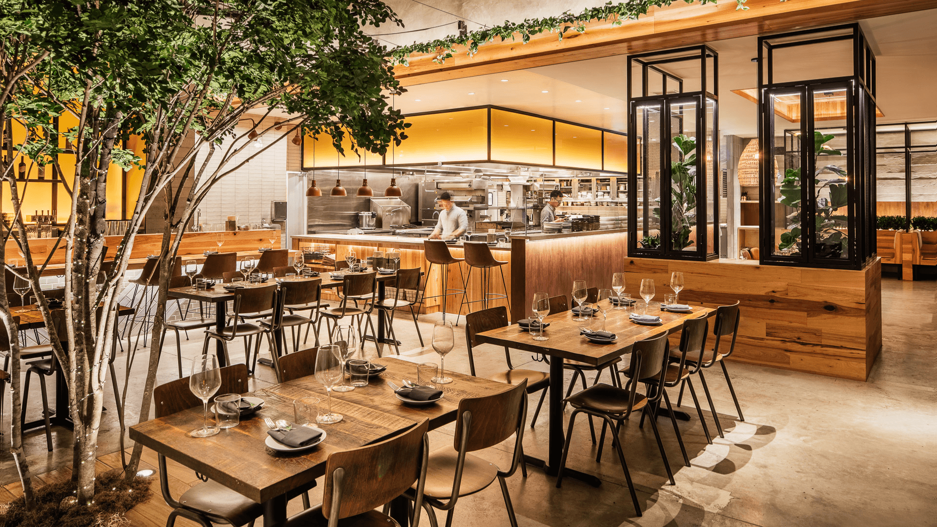 Interior of NEP Cafe’s Fountain Valley location featuring a warm modern dining space with wood accents, open kitchen views, lush greenery, and neatly set tables throughout the spacious restaurant.