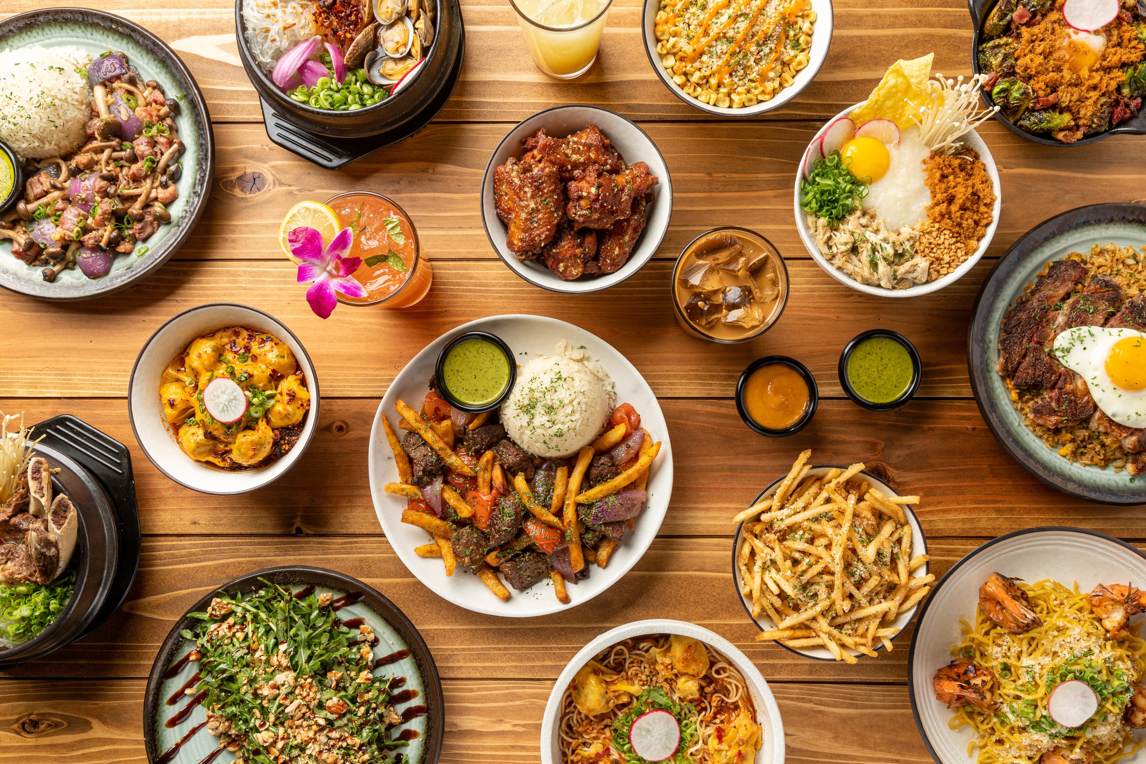 An overhead spread from VOX Kitchen featuring filet mignon lomo saltado with fries and rice, spicy wontons, parmesan garlic fries, crispy chicken wings, noodle dishes, and other signature Asian fusion plates arranged across a wooden table.