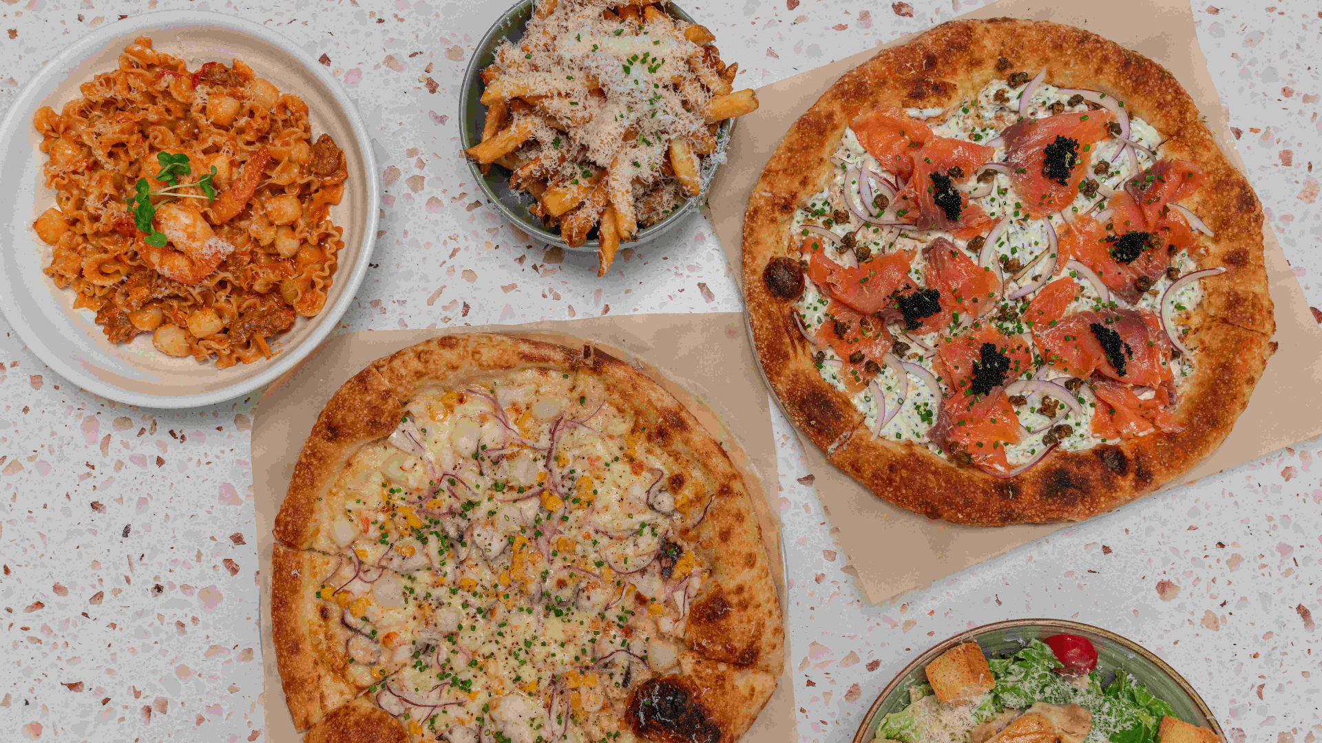 An overhead spread from INI Ristorante featuring mentaiko pizza topped with salmon and red onion, seafood mafaldine pasta in creamy tomato sauce, parmesan fries, Caesar salad, and assorted Italian-inspired dishes arranged on a terrazzo table.