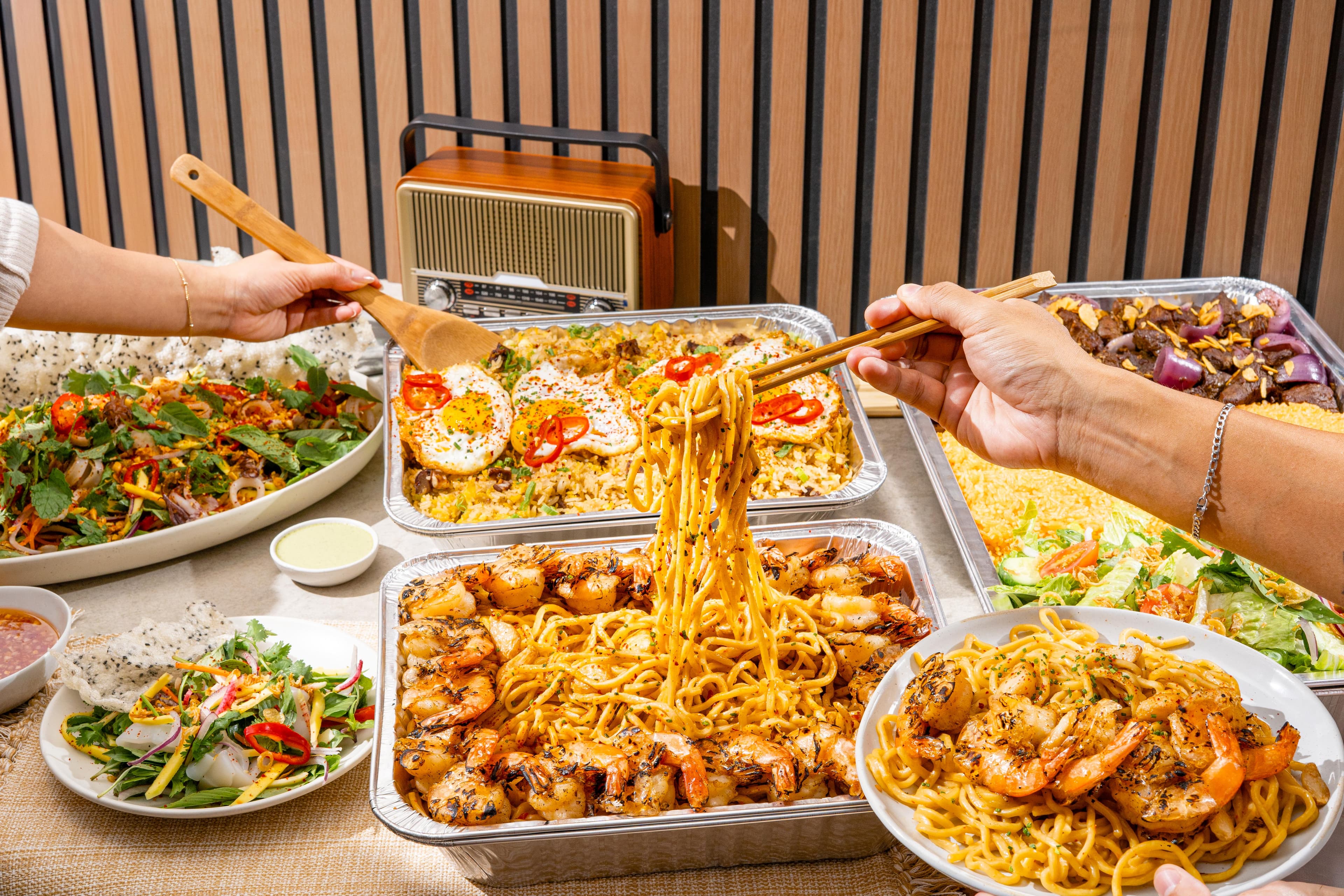 NEP Cafe party tray spread featuring garlic noodles with grilled shrimp, beef tongue fried rice topped with sunny-side eggs, grilled steak with rice, fresh salads, and Vietnamese-inspired catering trays, with guests serving and lifting noodles from the dishes.