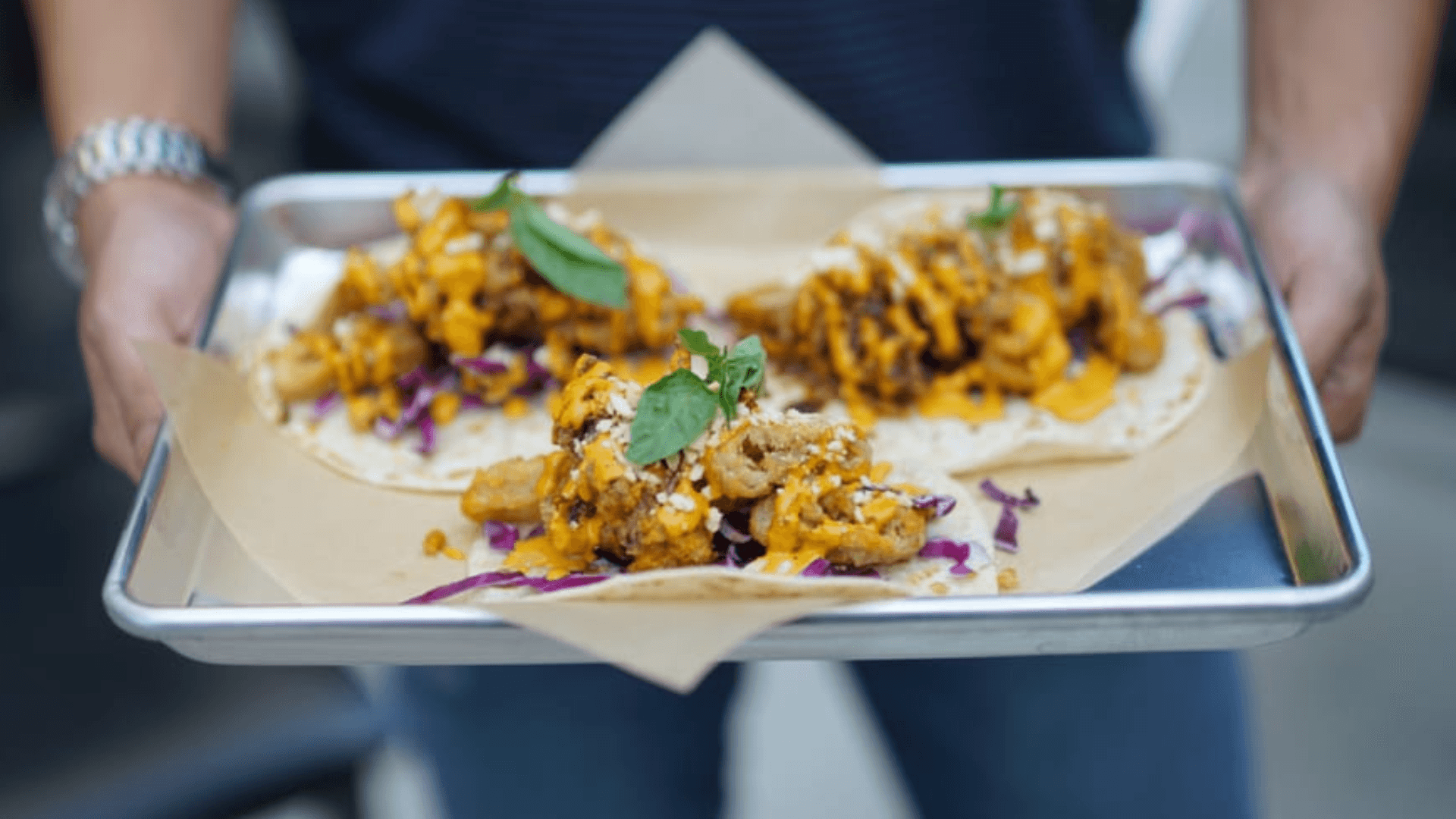 Close-up of Pango Taqueria tacos served on a metal tray, featuring crispy fried fillings topped with creamy orange sauce, fresh herbs, and shredded cabbage, held by a guest in an outdoor setting.