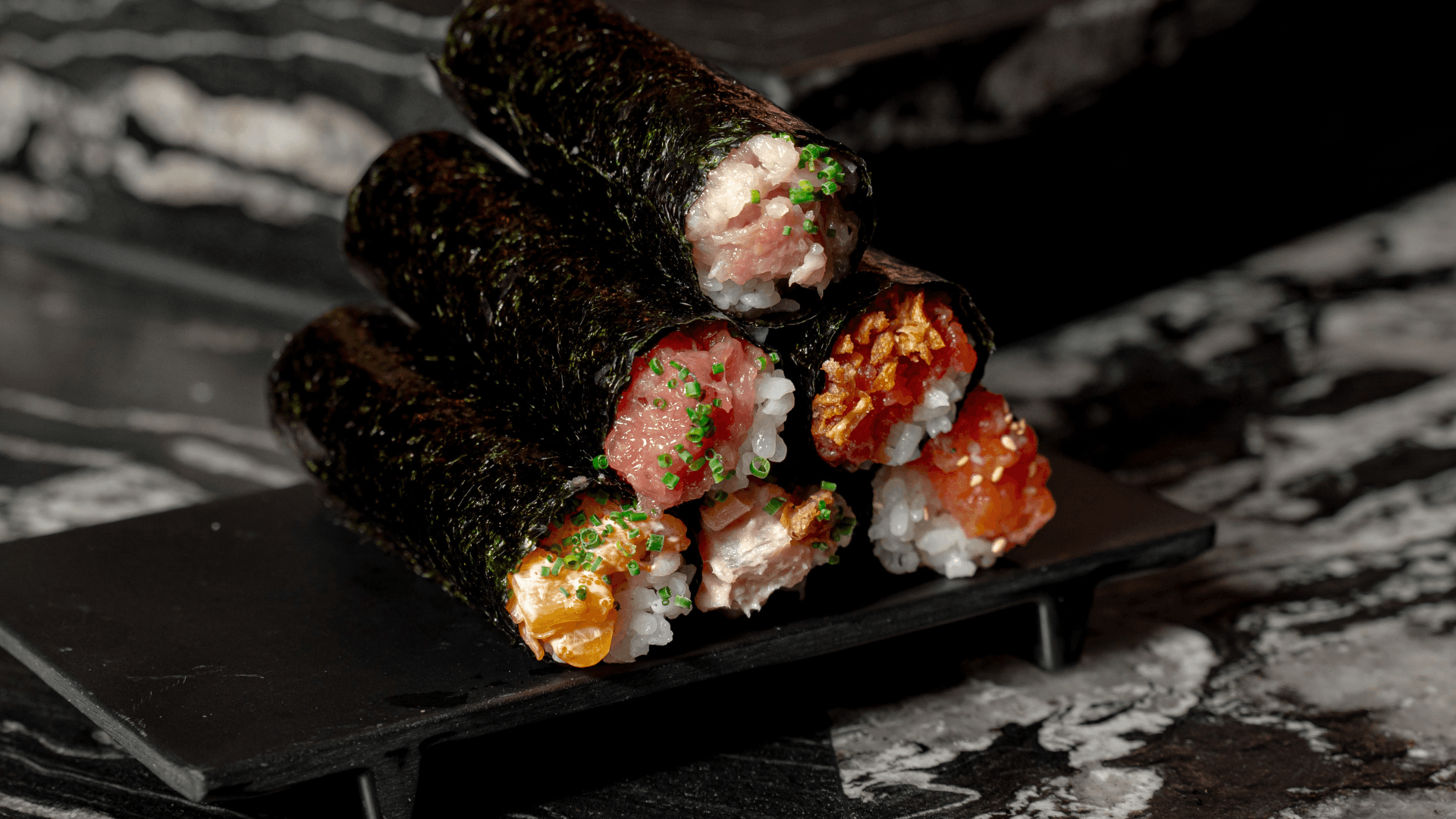 Stacked assortment of hand rolls from ROL Hand Roll Bar featuring fresh fish, crispy toppings, and sushi rice wrapped in seaweed, served on a black platter against a dark marble backdrop.