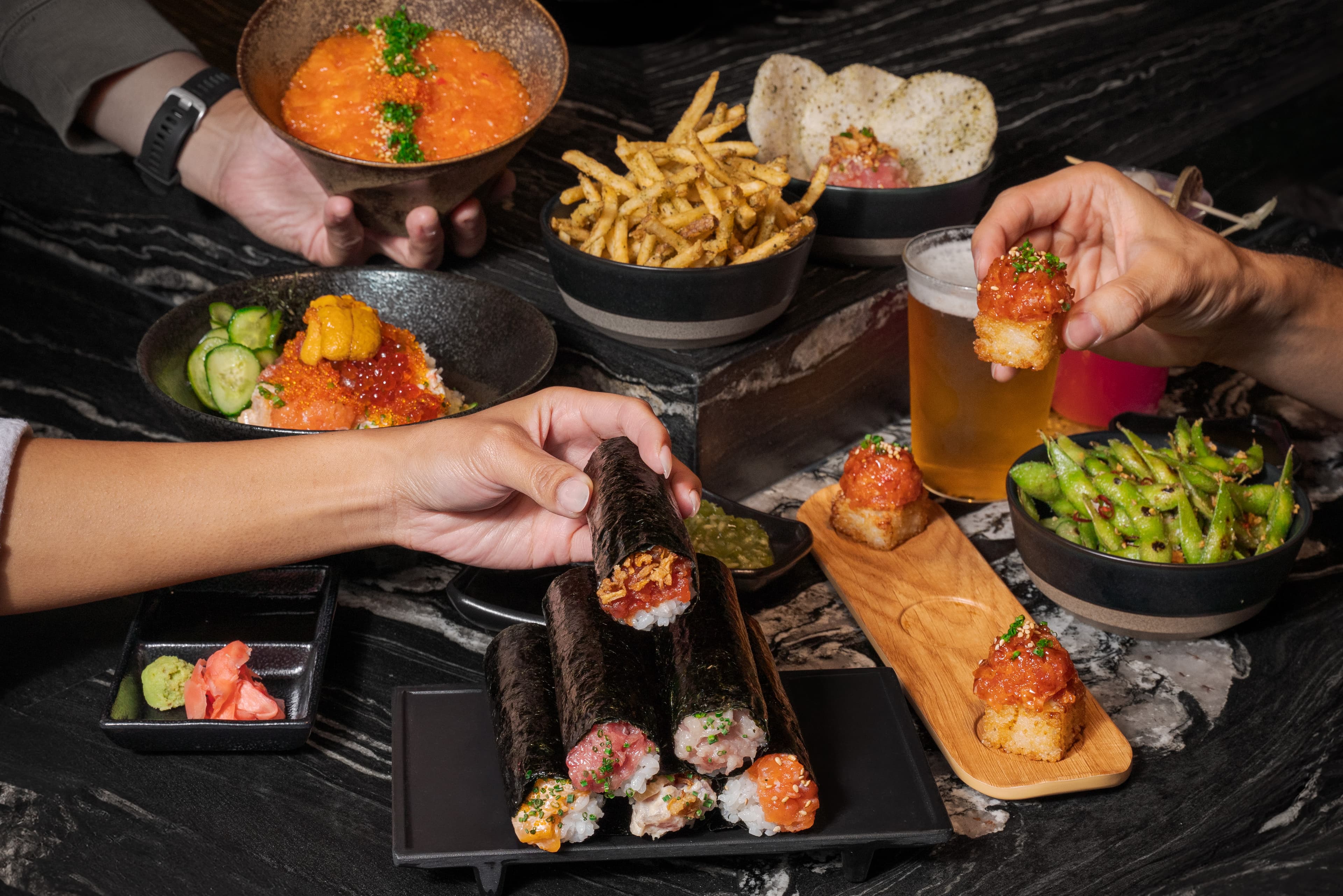Guests reaching for hand rolls and spicy tuna crispy rice at ROL Hand Roll Bar, surrounded by kaisendon, spicy edamame, seasoned fries, drinks, and Japanese-inspired small plates on a dark marble table.