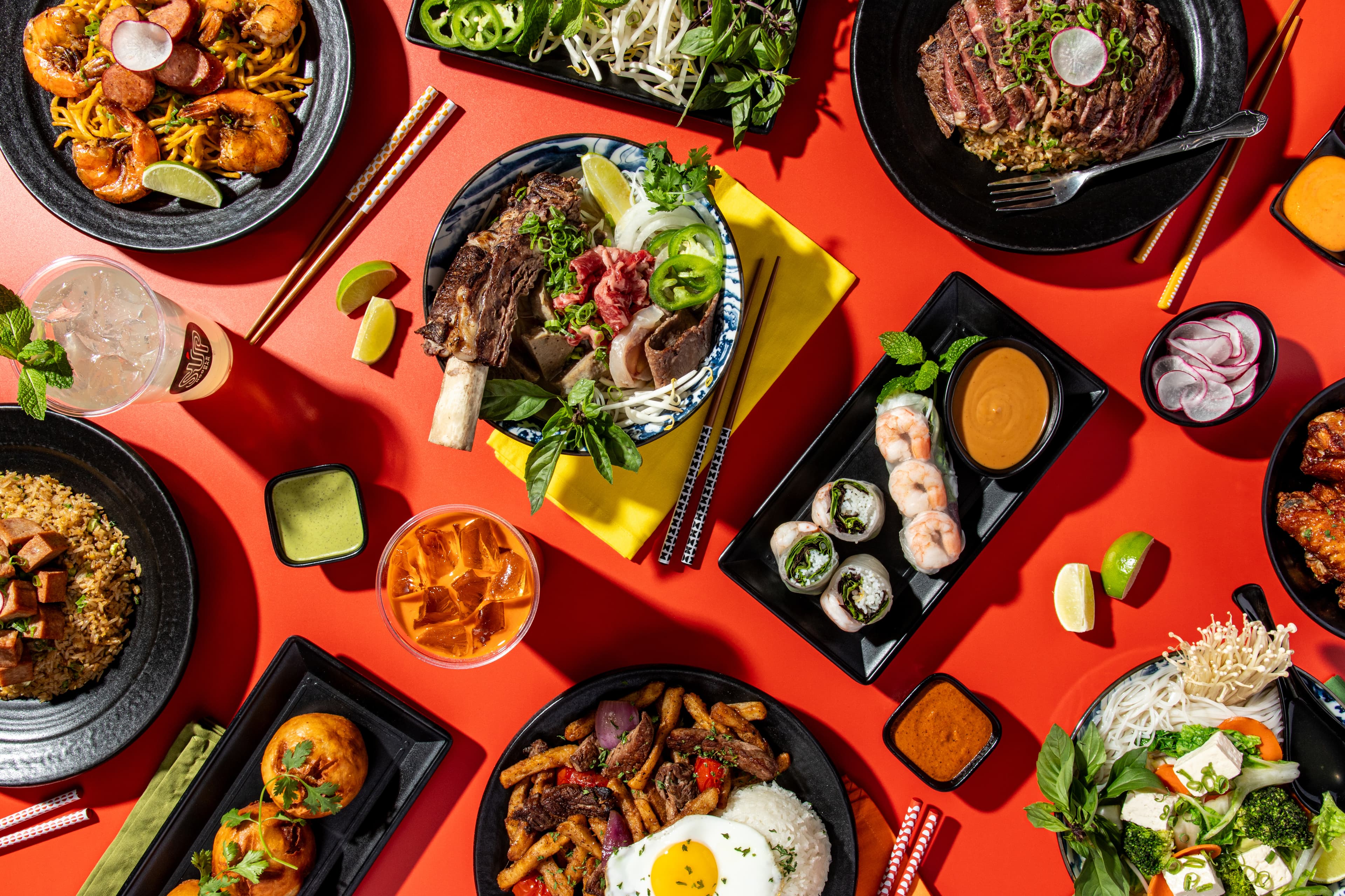 An overhead spread from SUP Noodle Bar featuring combination pho with fresh herbs, shrimp spring rolls with dipping sauce, savory lomo saltado, spam fried rice, and other Vietnamese-inspired favorites arranged across a bright red table.