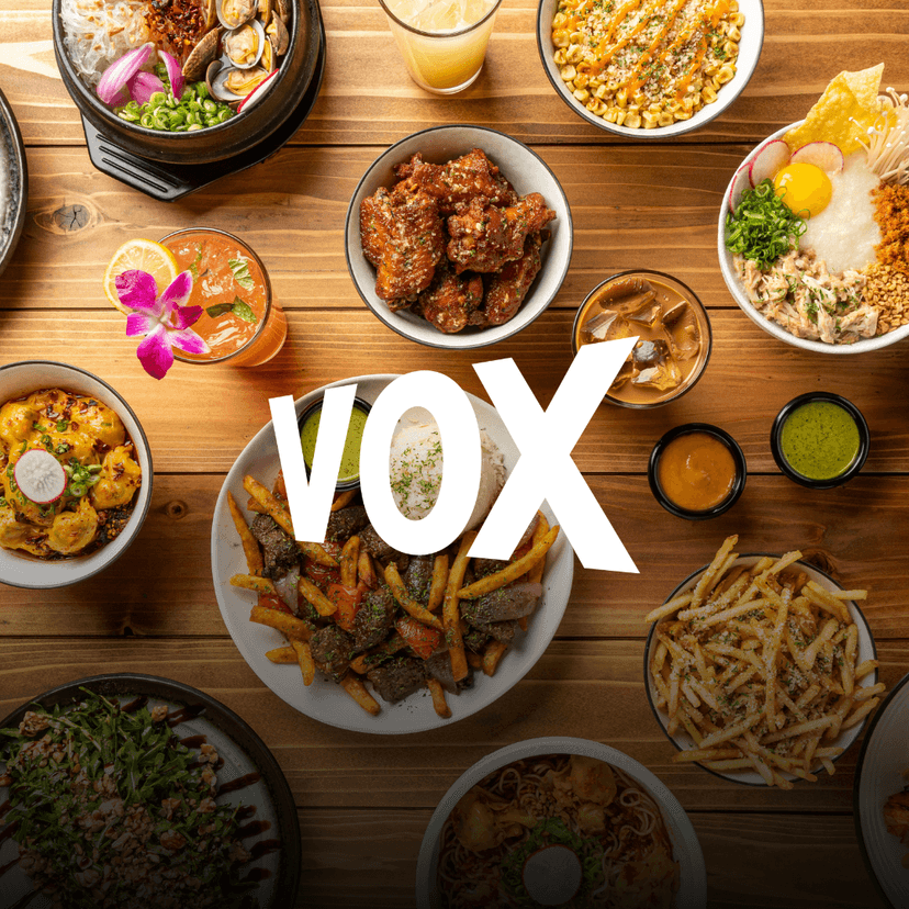 VOX Kitchen logo overlaid on an overhead spread from VOX Kitchen featuring filet mignon lomo saltado with fries and rice, spicy wontons, parmesan garlic fries, crispy chicken wings, noodle dishes, and other signature Asian fusion plates arranged across a wooden table.